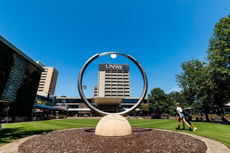Large circular metal sculpture with a sphere base in an outdoor campus setting, with UNSW building visible behind it and green trees framing the scene