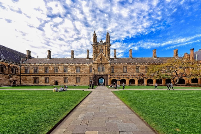 Historic stone university quadrangle with Gothic architecture, manicured green lawn, and central pathway under blue sky