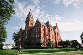 Historic red brick castle-style building with multiple turrets and towers set on green lawn under blue sky with clouds