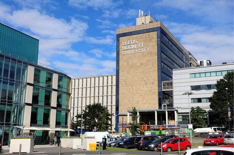 Modern Leeds city center buildings with green glass facade, tall brick tower, and parked cars under blue sky